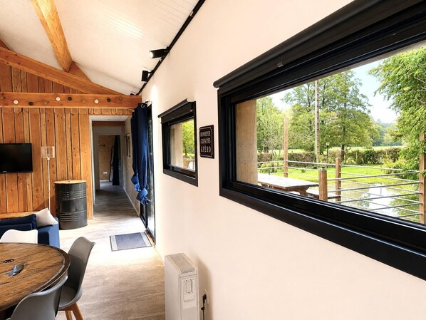 A room with a wooden ceiling, a dining table with chairs, a TV mounted on the wall, and a window with a view of a garden.
