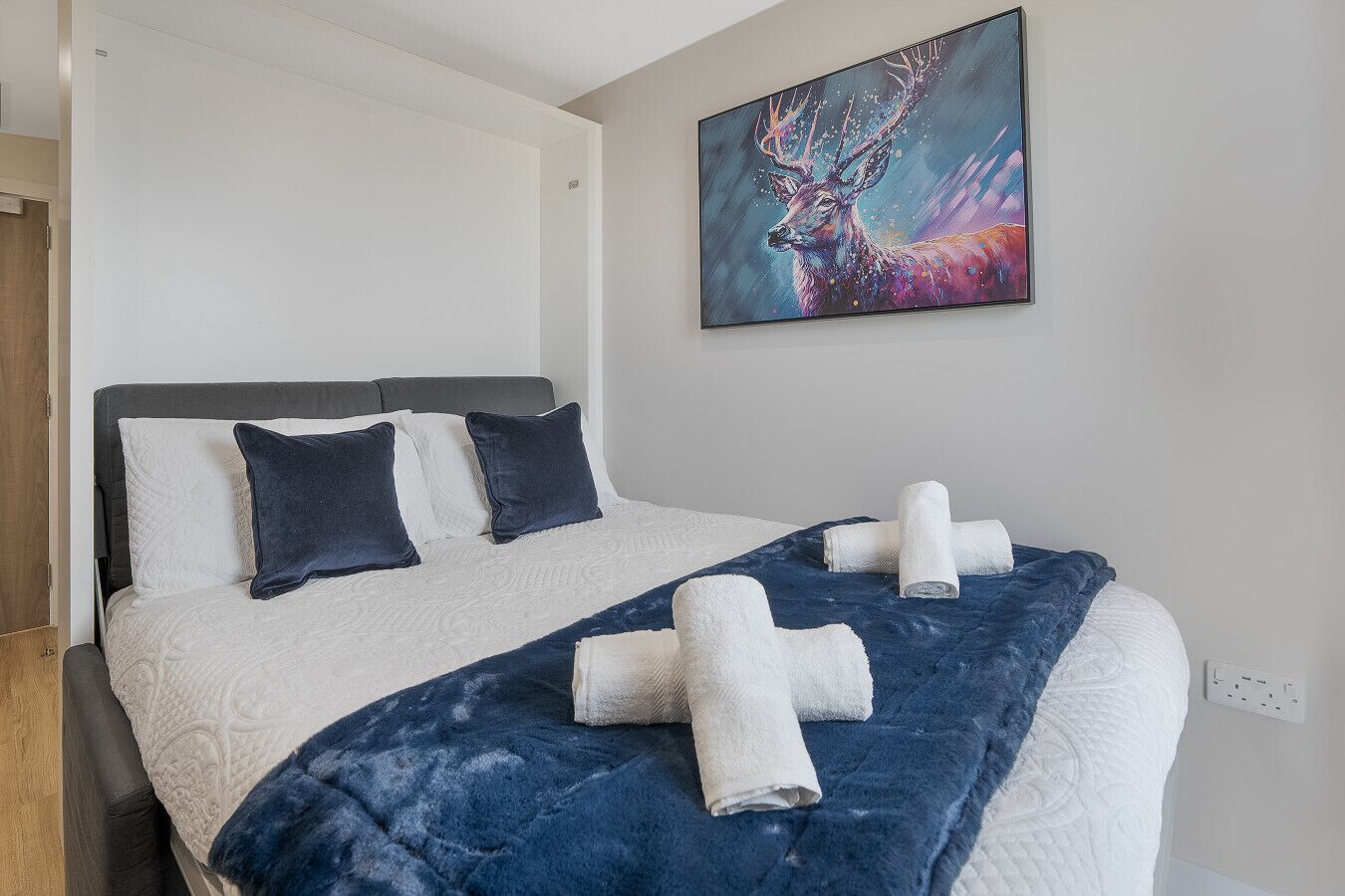 Un lit bien fait, avec une couverture bleue et des serviettes blanches, une tête de lit grise et un tableau représentant un cerf accroché au mur.