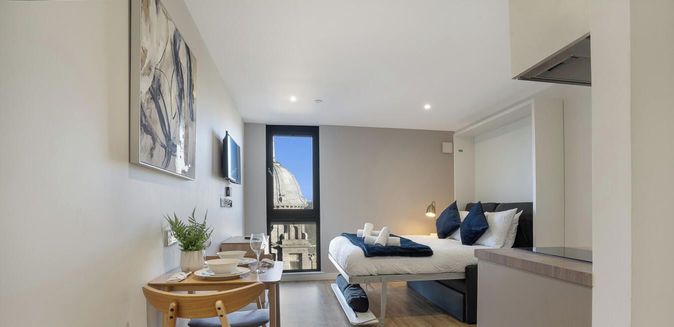 Quarto moderno com cama, mesa de cabeceira e vista para um edifício através de uma janela.
