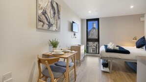 Una habitación de hotel moderna con área de comedor, una cama con vista a un edificio y un cuadro en la pared.