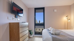A modern hotel room with a large flat-screen TV displaying 'NETFLIX', a wooden dresser, a potted plant, a bed with neatly folded towels, and a view of a historic building through a window. - The Point 925 - Grampian Lettings (Aberdeen)