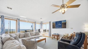A spacious living room with a large sofa, a recliner, and a coffee table. There is a ceiling fan, a framed picture on the wall, and large windows offering a view of the landscape. - Babe's Place | 105 ft to the beach | Dog Friendly (Kill Devil Hills)
