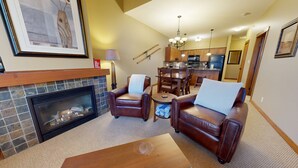 Ein Wohnzimmer mit Kamin, Ledersofas, einem Couchtisch und einem Essbereich mit Stühlen, im Hintergrund befindet sich eine Küche.