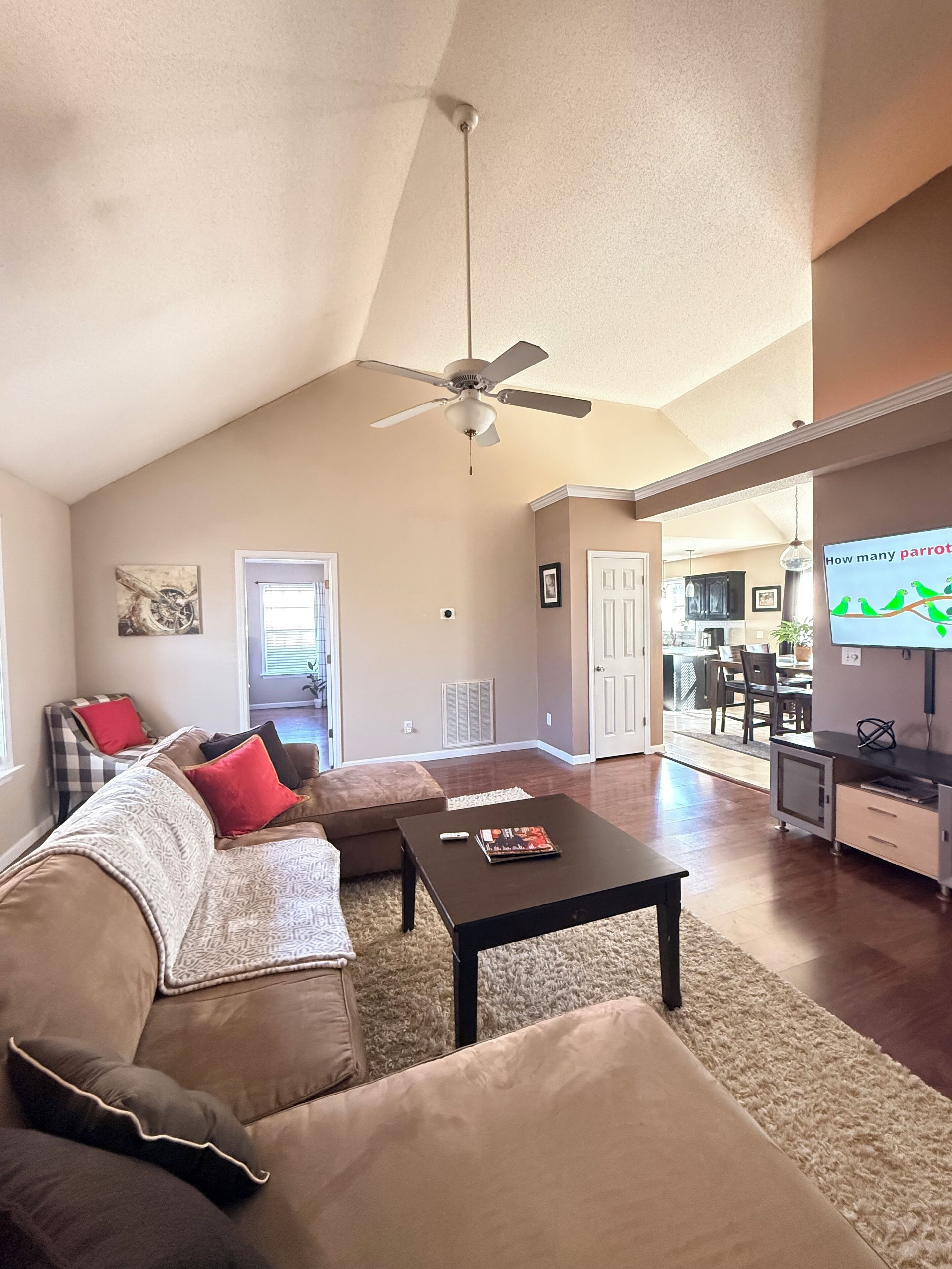 A spacious living room with a high ceiling, a ceiling fan, a sofa with cushions, a coffee table, and a television.