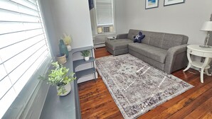 Uma sala de estar com um sofá cinza, um tapete estampado e uma pequena mesa branca com um abajur.