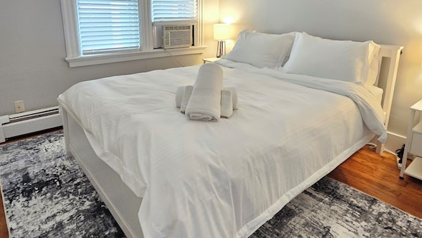 Cama bem arrumada com lençóis brancos, tapete com padrão preto e branco, mesa de cabeceira com abajur e janela com persianas.
