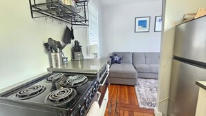 Cozinha compacta com fogão, forno e micro-ondas integrado. Há um sofá cinza e um tapete pequeno na sala.