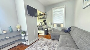 Uma sala de estar moderna com um sofá cinzento, uma TV de tela plana na parede, uma área de cozinha ao fundo e um tapete estampado no chão.