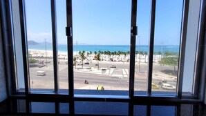 A view through a window with vertical blinds, overlooking a beach with palm trees and a clear blue sky. - Copacabana Oceanfront • 3 Spacious Bedrooms • Incredible Ocean View (Rio de Janeiro)