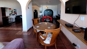 A dining area with a round table and chairs, a kitchen in the background, and a TV mounted on the wall. - Copacabana Oceanfront • 3 Spacious Bedrooms • Incredible Ocean View (Rio de Janeiro)