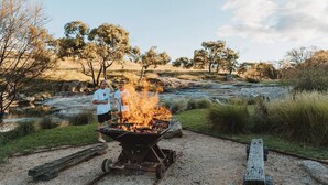 A campfire with two people nearby, set in a natural outdoor environment with trees and a river in the background.