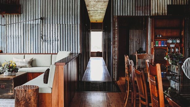 A rustic interior with a wooden floor, a sofa, a bar area with chairs, and a corrugated metal wall.