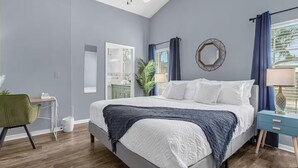 Une chambre à coucher avec un lit, un bureau, une chaise, un miroir et une fenêtre avec des rideaux.