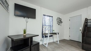 A modern living room with a flat-screen TV mounted on the wall, a dark wooden coffee table with a plant, a glass-top side table, and a staircase leading to a door. - Chill Studio Pensacola Getaway (Pensacola)