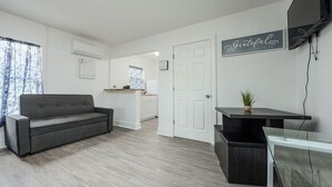 A modern living room with a grey sofa, a small kitchen area, and a TV mounted on the wall. - Chill Studio Pensacola Getaway (Pensacola)