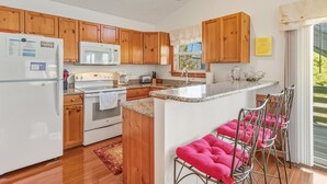 Una cocina con gabinetes de madera, un refrigerador blanco, un microondas, una estufa y un lavamanos. Hay taburetes de bar color rosa y una ventana con vista a follaje.