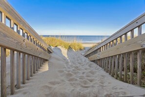 Une promenade en bois qui mène à une plage bordée de sable et d’herbe de chaque côté.