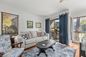 A living room with a sofa, armchair, coffee table, and a view through a glass door to the outside. - Studio Seven at Pine Valley (Pinehurst)