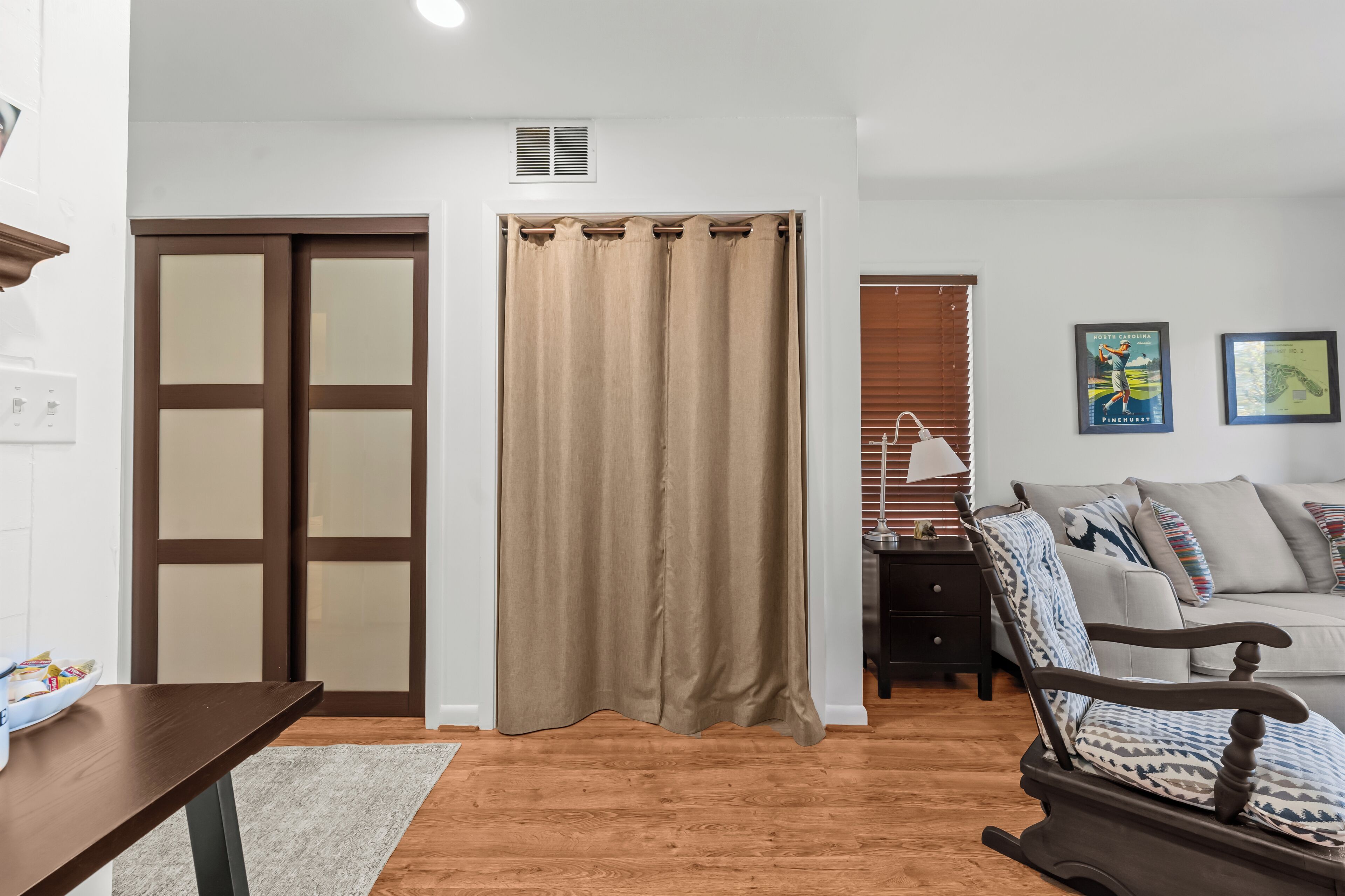 A living room with a brown wooden floor, a brown curtain, a sofa, a rocking chair, and a small table.