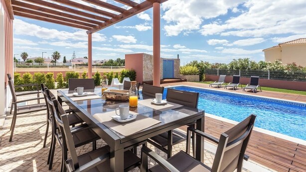 A poolside area with a table set for breakfast, a pool, and a pergola structure. - Blue Heaven Luxury V5 (Porches)