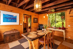 Interior de madera con una mesa de comedor, sillas, una araña, obras de arte y un ventanal con follaje exterior.