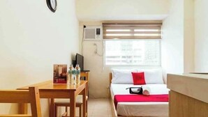 Une petite pièce propre, avec un lit, un bureau et une chaise. Il y a une fenêtre avec des stores, un climatiseur et une horloge au mur.