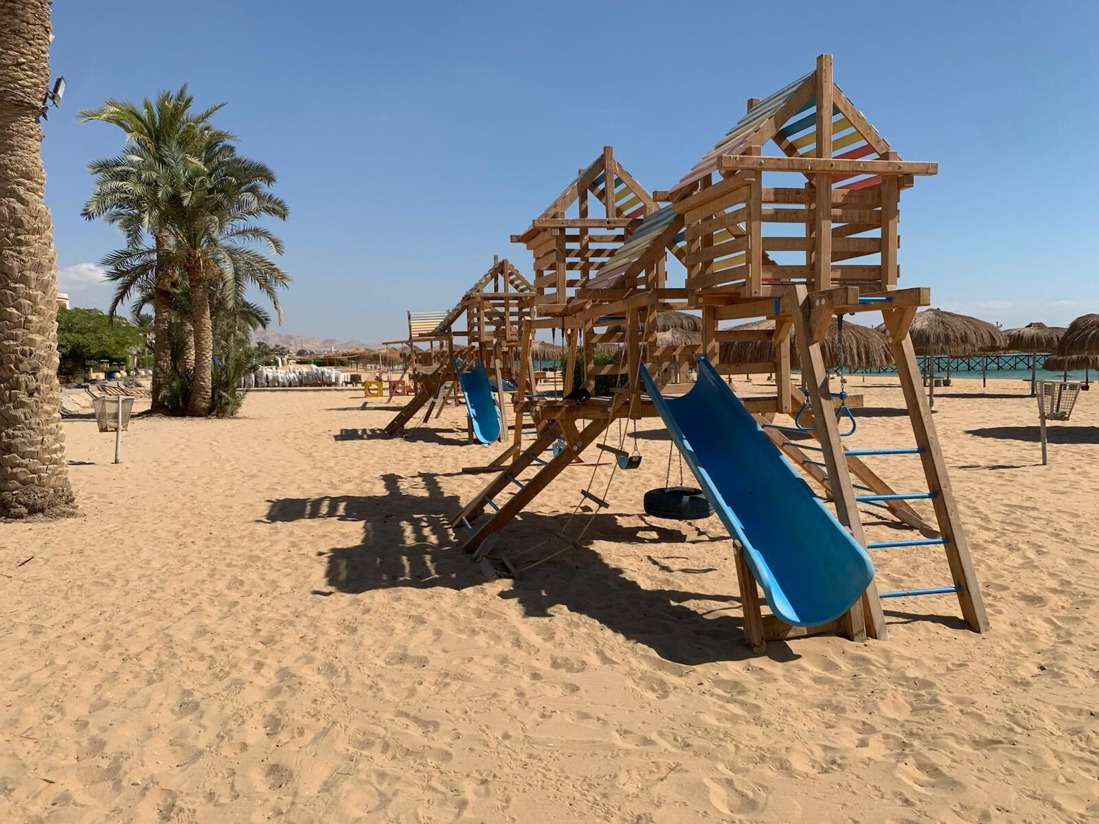 A sandy beach with wooden play structures, including slides and swings, and palm trees in the background.