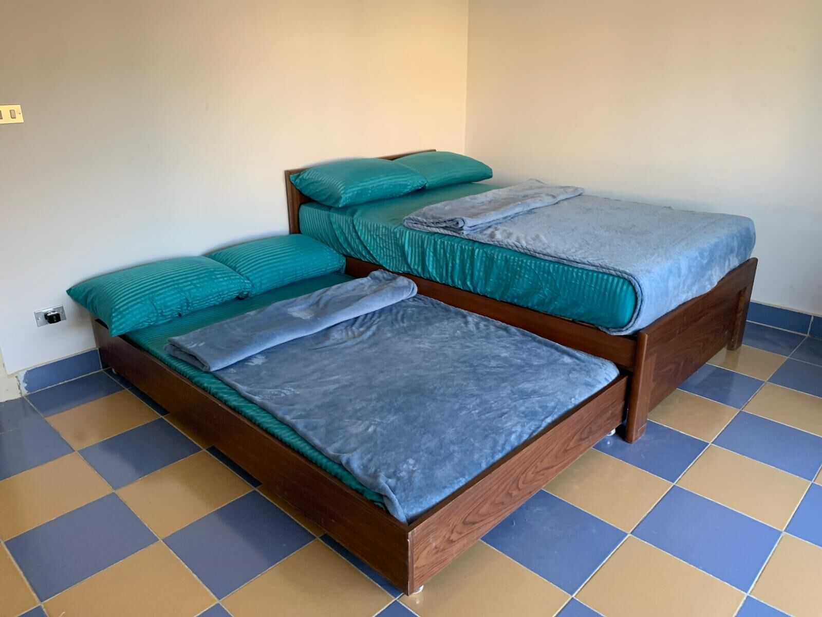 A room with two wooden beds, each with teal bedding and a grey blanket. The floor has a blue and beige checkered pattern.