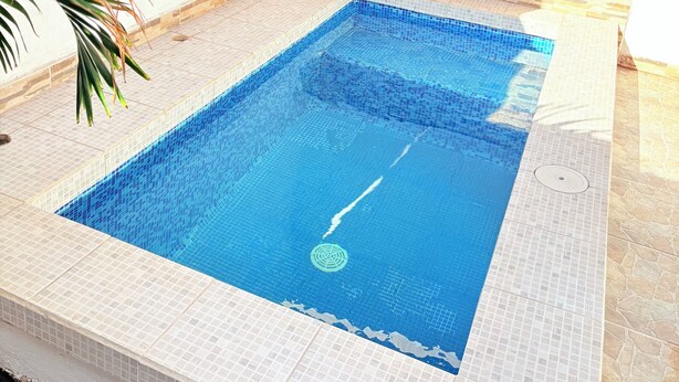 A rectangular swimming pool with blue tiles and a white border, surrounded by a tiled deck and a stone wall.