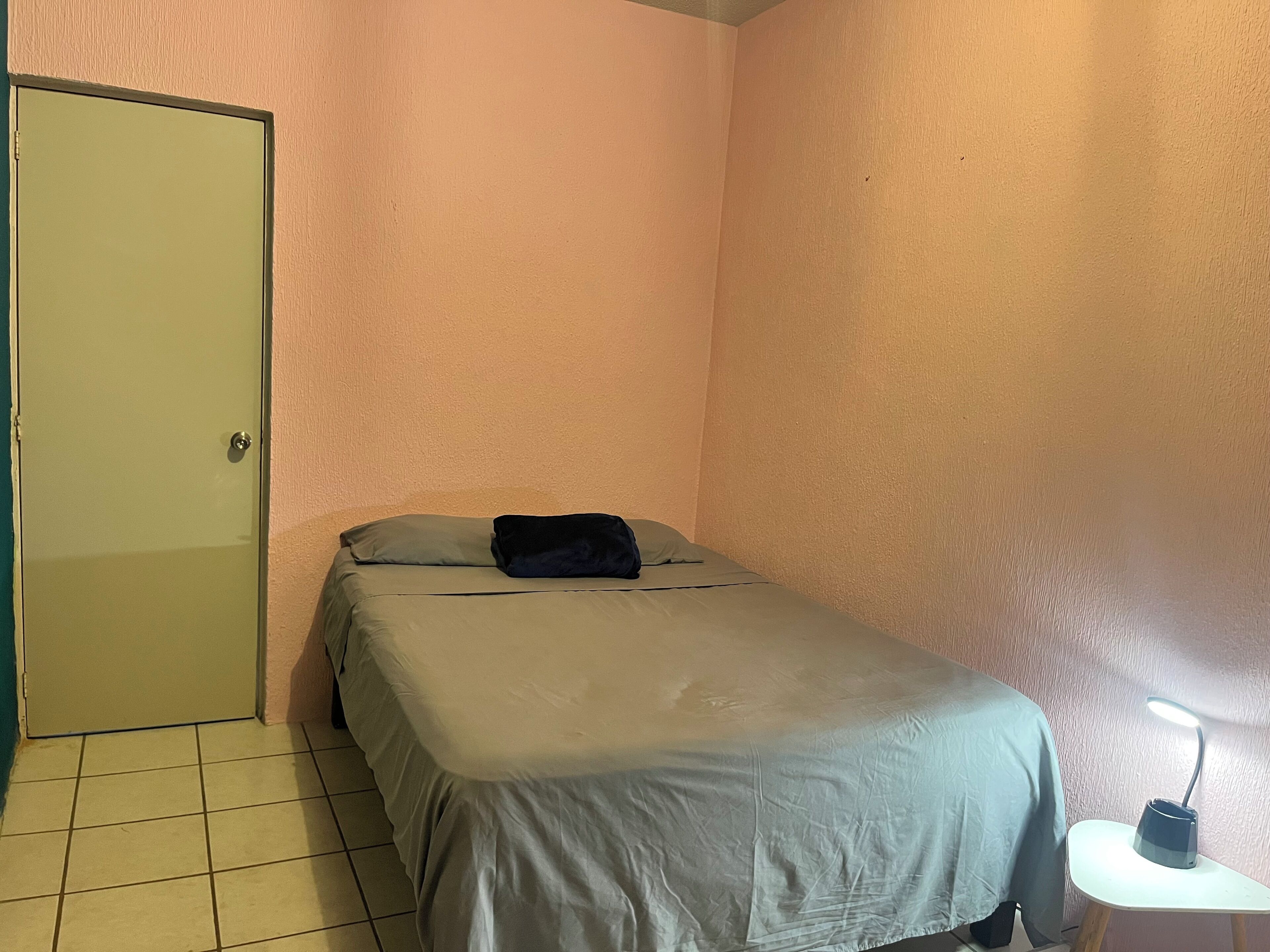 A small, single-bed room with a green door, a bedside table with a lamp, and a plain wall.
