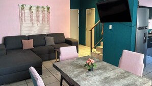 A modern living room with a sofa, a dining table with a potted plant, and a kitchen area with a refrigerator.