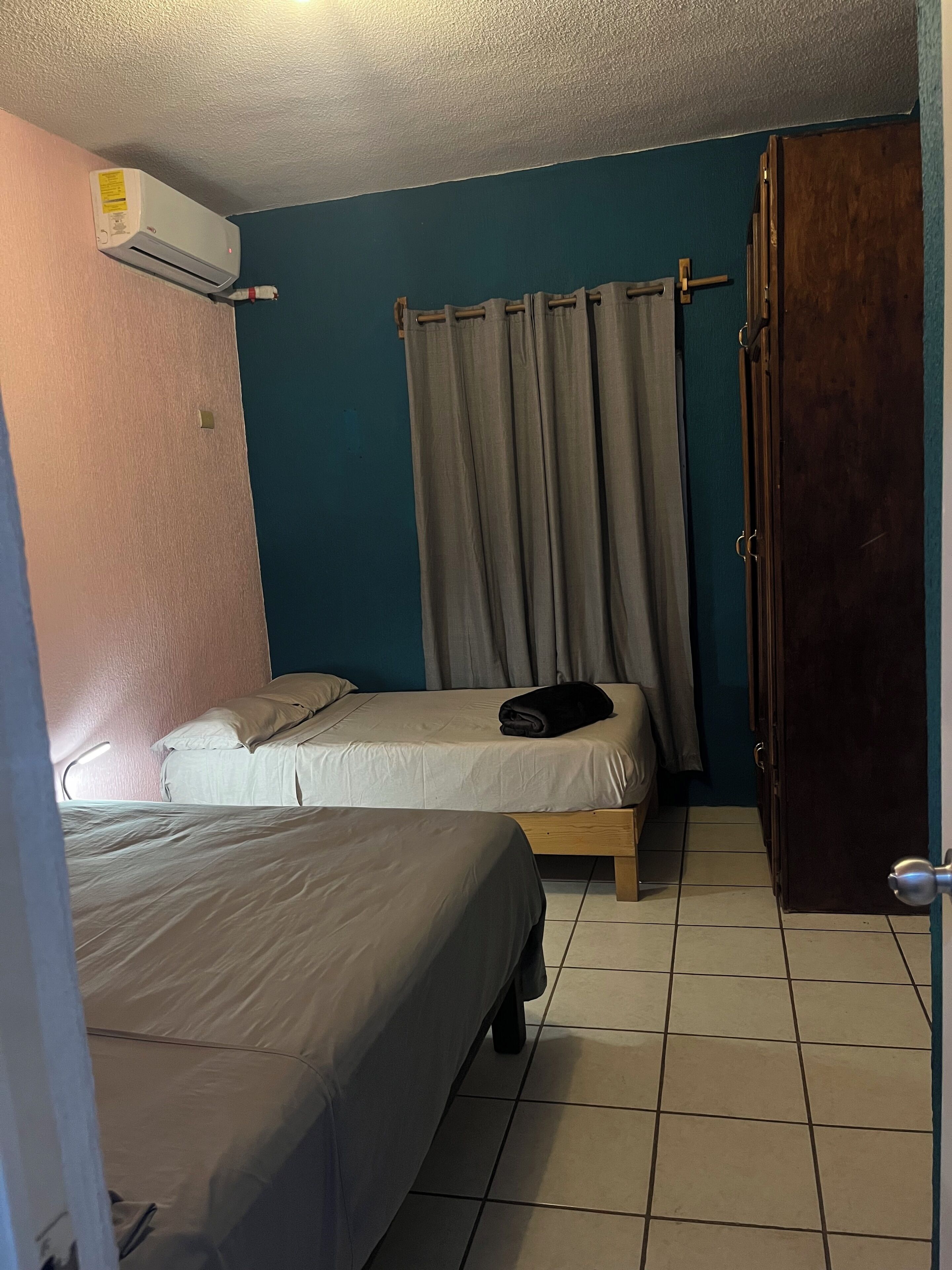 A small, compact room with two single beds, a wooden wardrobe, a curtain, and an air conditioning unit mounted on the wall.