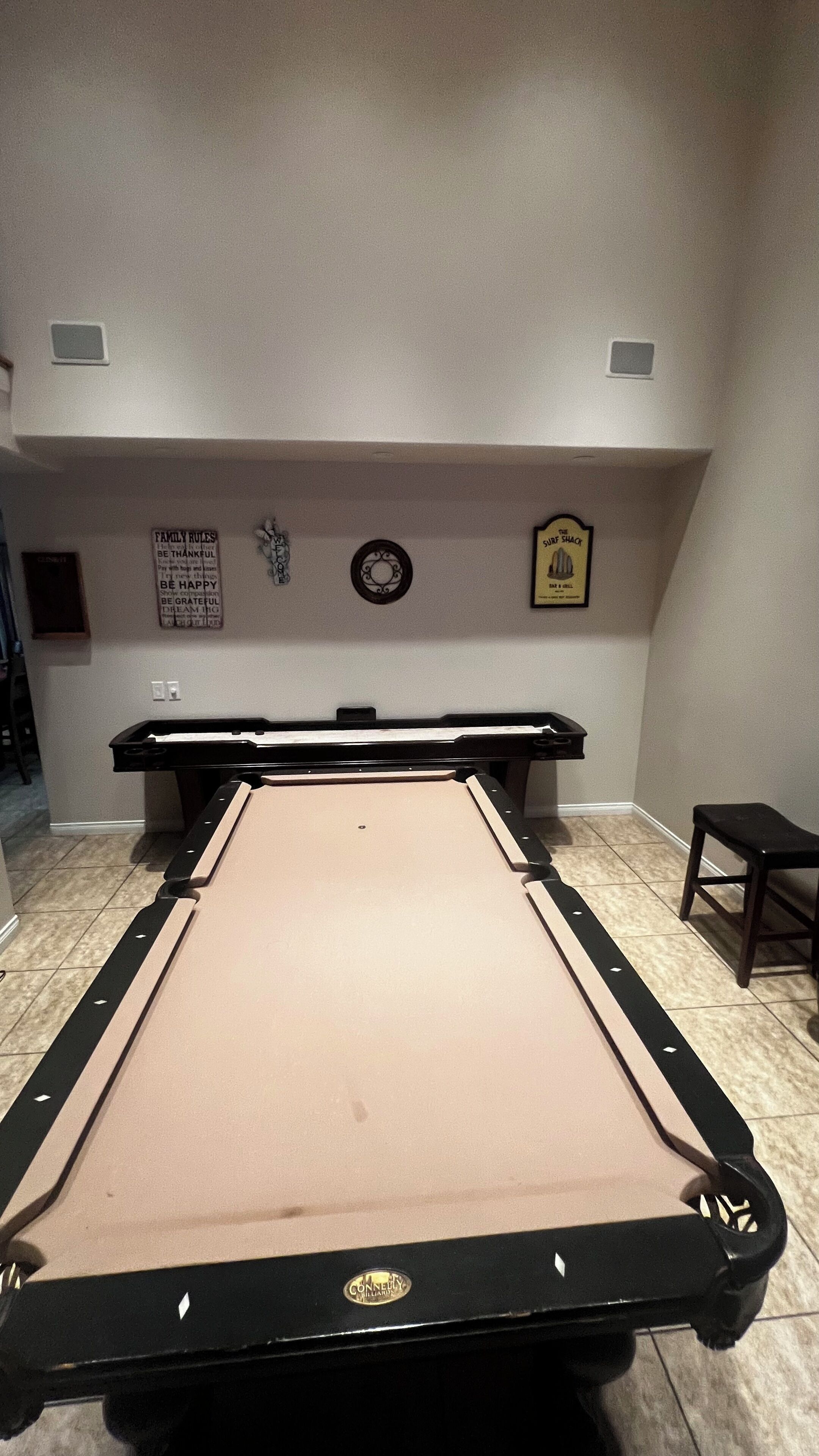 Uma mesa de bilhar com superfície feltro bege e bordas pretas, cercada por um piso ladrilhado.