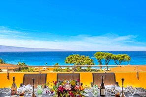 A dining table set with chairs, a bottle of wine, and a vase of flowers, overlooking a beach and ocean. - Maui Kaanapali | Penthouse 2 BR | Car Incl. w/ 6+ Nights | MKV-A606 by KBM (Lahaina)