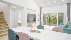 A modern living room with a white dining table, teal and pink chairs, a flat-screen TV, and a large window with a view of trees. - Gangneung Pinehill Park Pension&caravan (Gangneung)