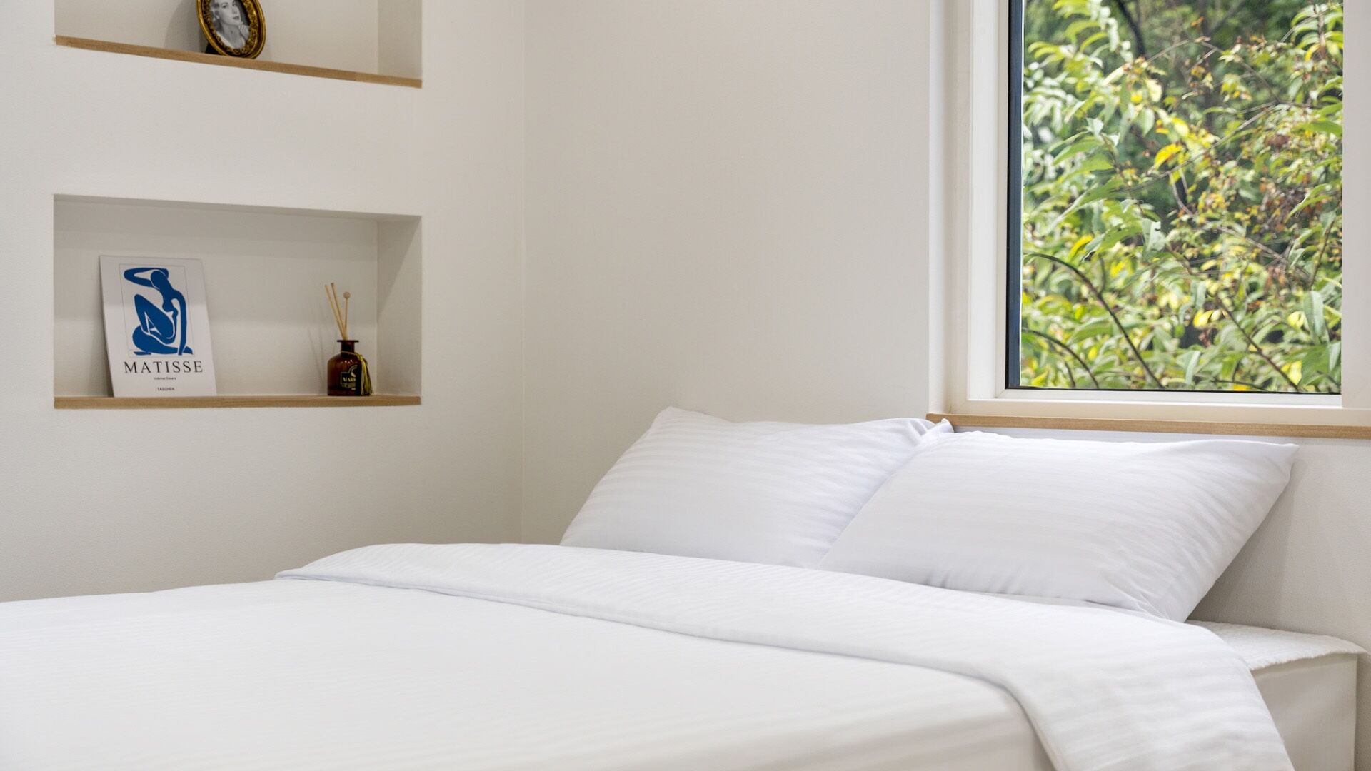 A bed with white bedding, a window with a view of greenery, and a shelf with a framed picture and a bottle.