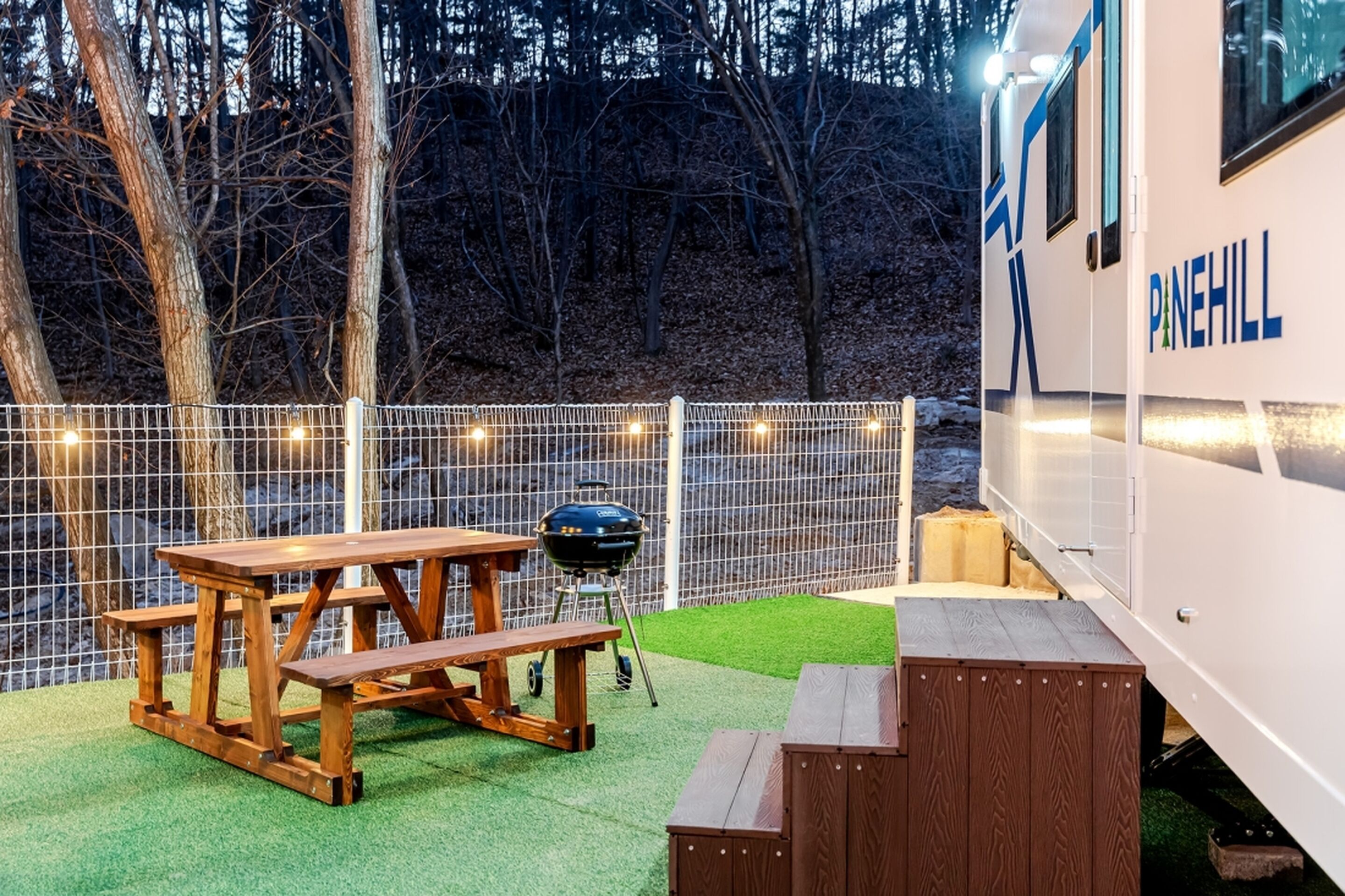 Un camping-car Pinehill équipé d’une table de pique-nique et d’un barbecue, entouré d’une clôture blanche et illuminé de guirlandes lumineuses.