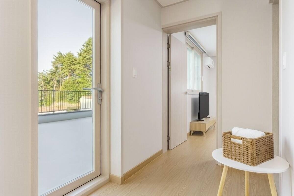 Un pasillo con una puerta corrediza de vidrio que da a un balcón, un televisor en una habitación y una cesta de mimbre sobre una mesita.