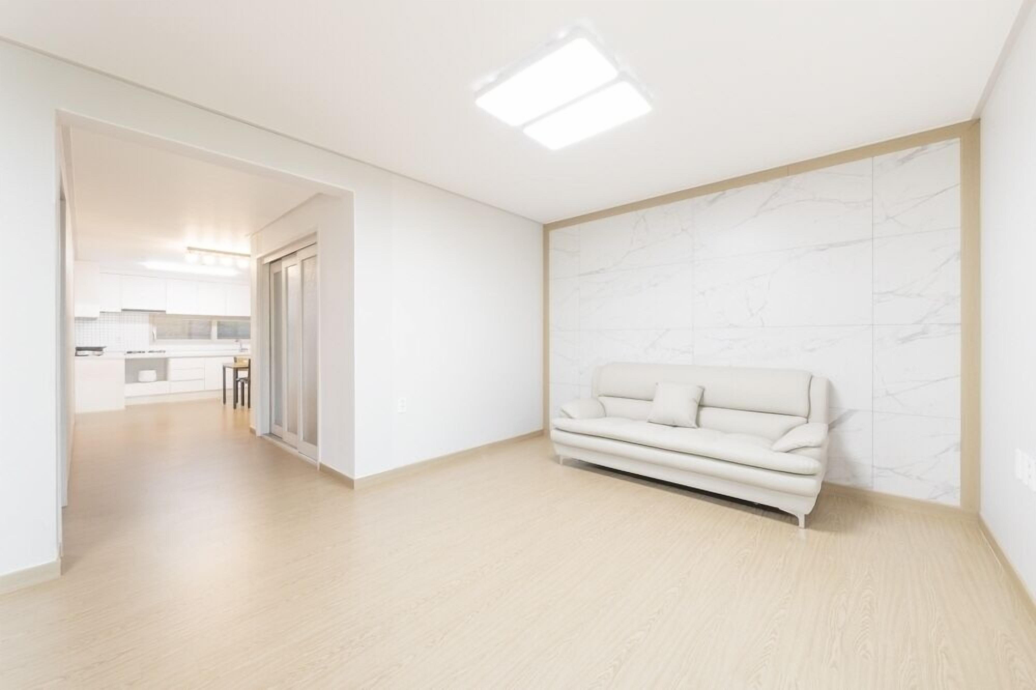 Um interior moderno com design minimalista, apresentando um sofá de cor clara em frente a uma parede de mármore, piso de madeira e uma área de cozinha visível ao fundo.