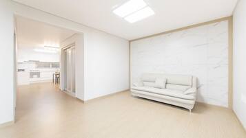 Um interior moderno com design minimalista, apresentando um sofá de cor clara em frente a uma parede de mármore, piso de madeira e uma área de cozinha visível ao fundo.