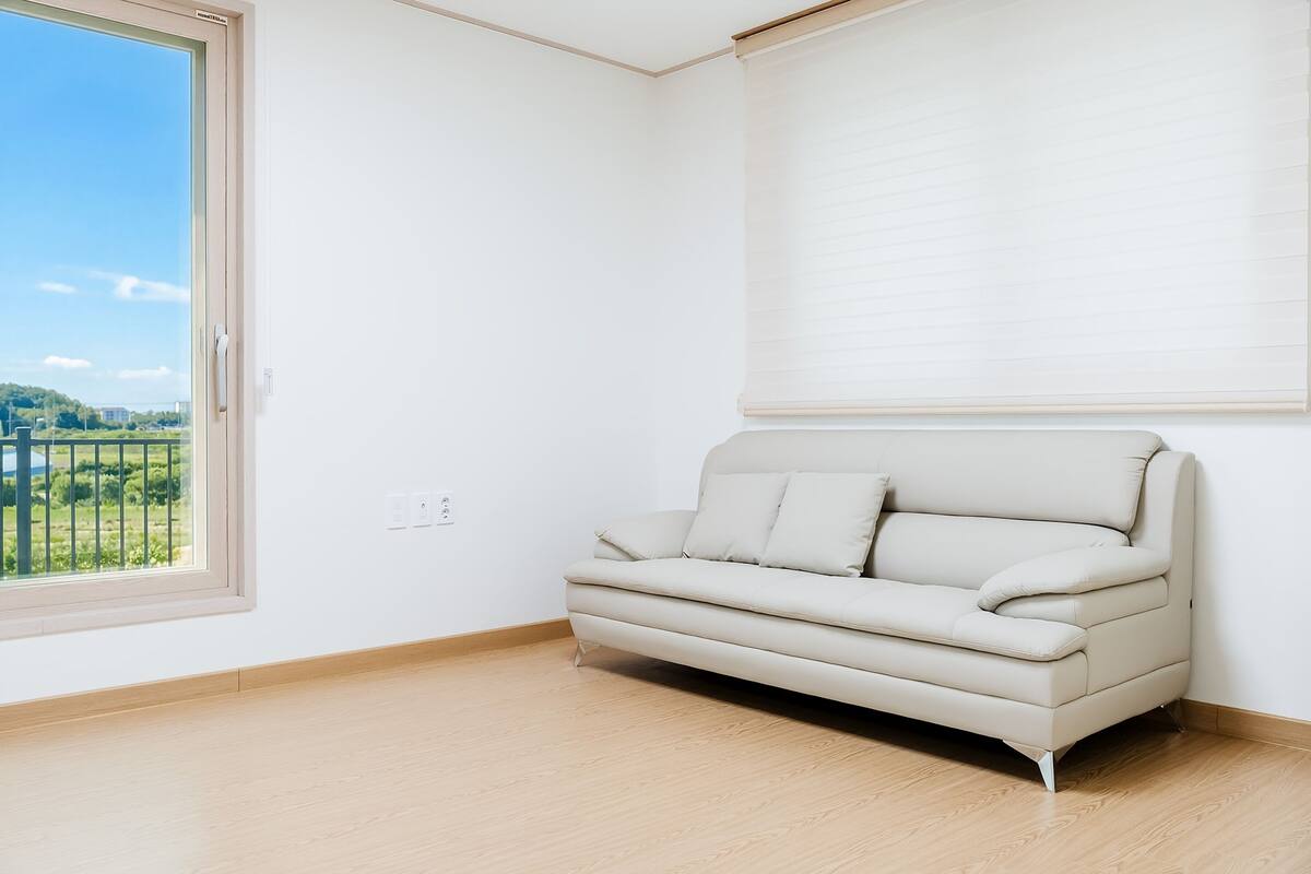 Una habitación con un sofá color beige, un ventanal con vistas a follaje, y una pared blanca.