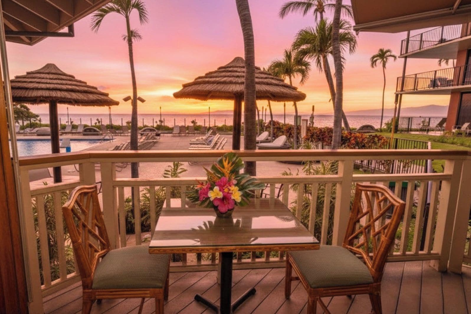 A balcony with a table and chairs, overlooking a pool and beach area with umbrellas and palm trees.