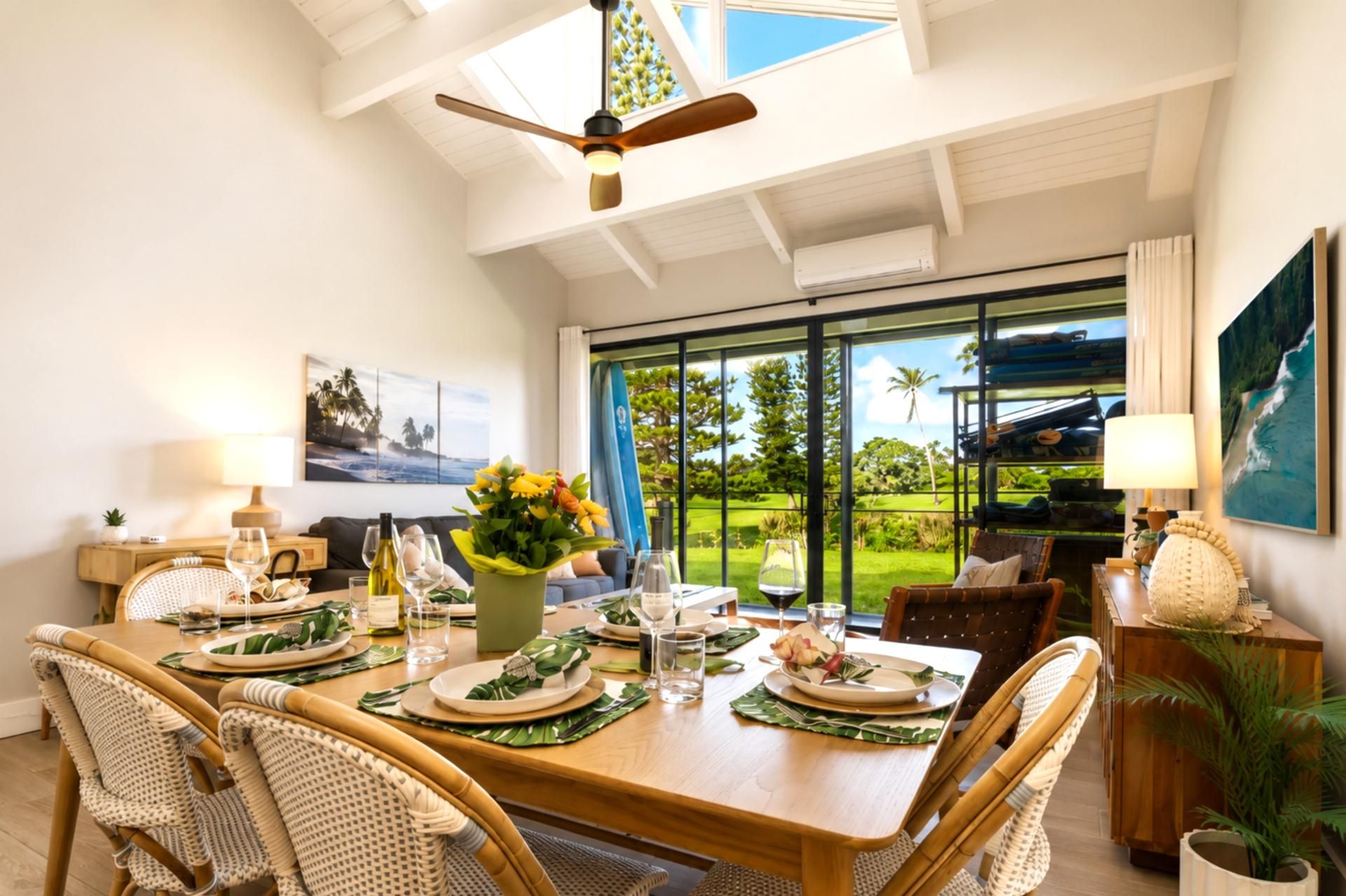 Un coin repas avec une table dressée pour quatre personnes, un ventilateur de plafond et une vue sur un jardin luxuriant, visible à travers de grandes fenêtres.