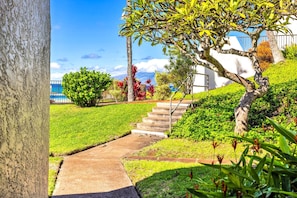 A pathway leading to a building with a white exterior, surrounded by lush greenery and a clear sky. - Napili Point | Oceanfront 2 BR, Sleeps 8 | Car Incl. w/ 6+ Nights | NAP ML-2490 by KBM (Lahaina)