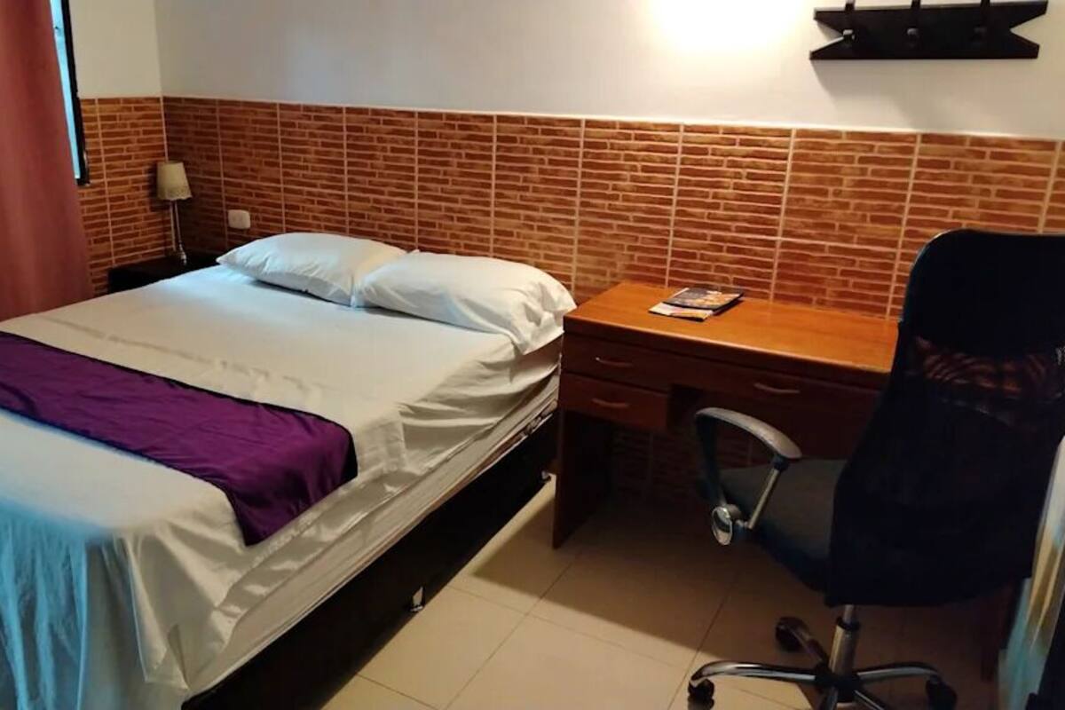 A hotel room with a bed, a desk, a chair, and a brick-patterned wall.