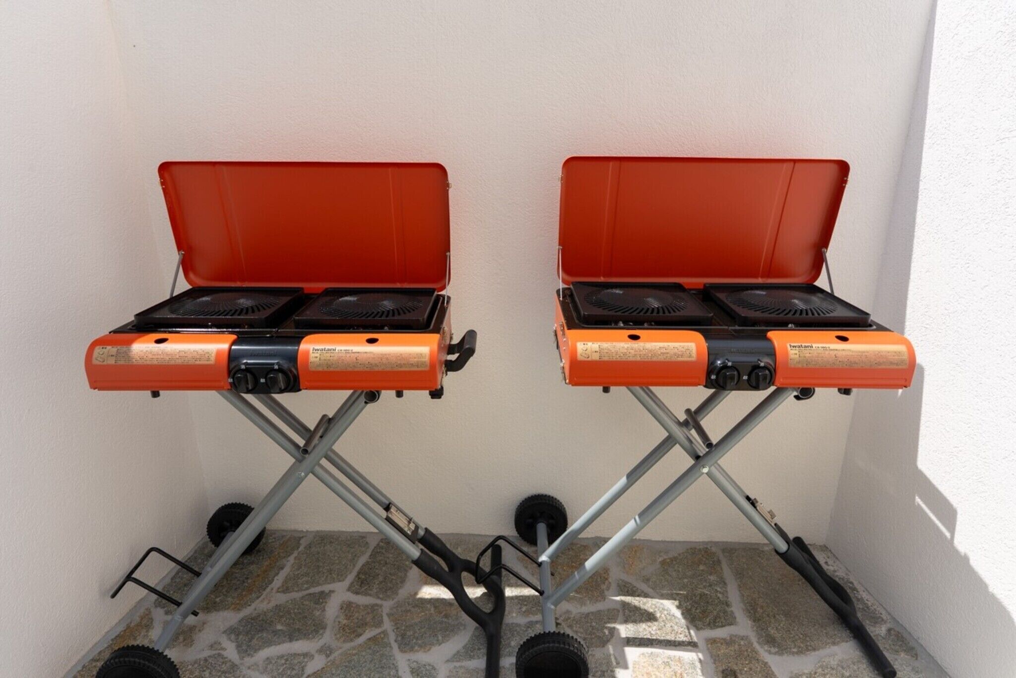 Deux réchauds à gaz portables, dotés de couvercles rouges et de grilles noires, sont fixés sur un cadre métallique muni de roues, le tout adossé à un mur blanc.