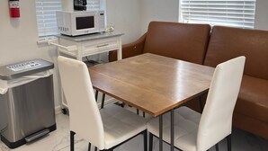 Uma sala de jantar com uma mesa de madeira e cadeiras brancas, micro-ondas, cafeteira e lixeira.