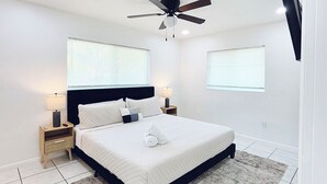 Ein Schlafzimmer mit einem Bett, Nachttischen, einem Deckenventilator und zwei Fenstern mit Jalousien.