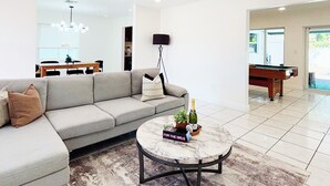 Ein modernes Wohnzimmer mit einem Ecksofa, einem runden Couchtisch mit Flasche und Buch, einem Billardtisch und einem Essbereich im Hintergrund.