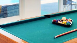 Ein Billardtisch mit Kugeln und Spielstange in einem Raum mit großen Fenstern, die auf einen Swimmingpool blicken.